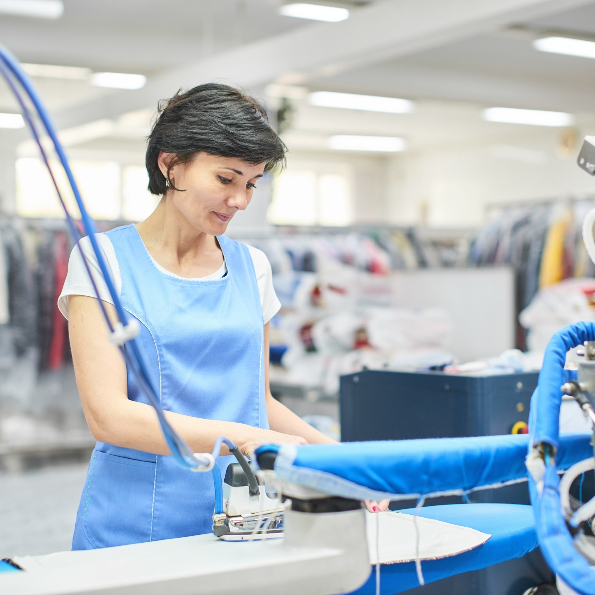 Woman in blue uniform steam irons clothes at a Inner West Sydney laundry with racks behind, convenient pickup and delivery.
